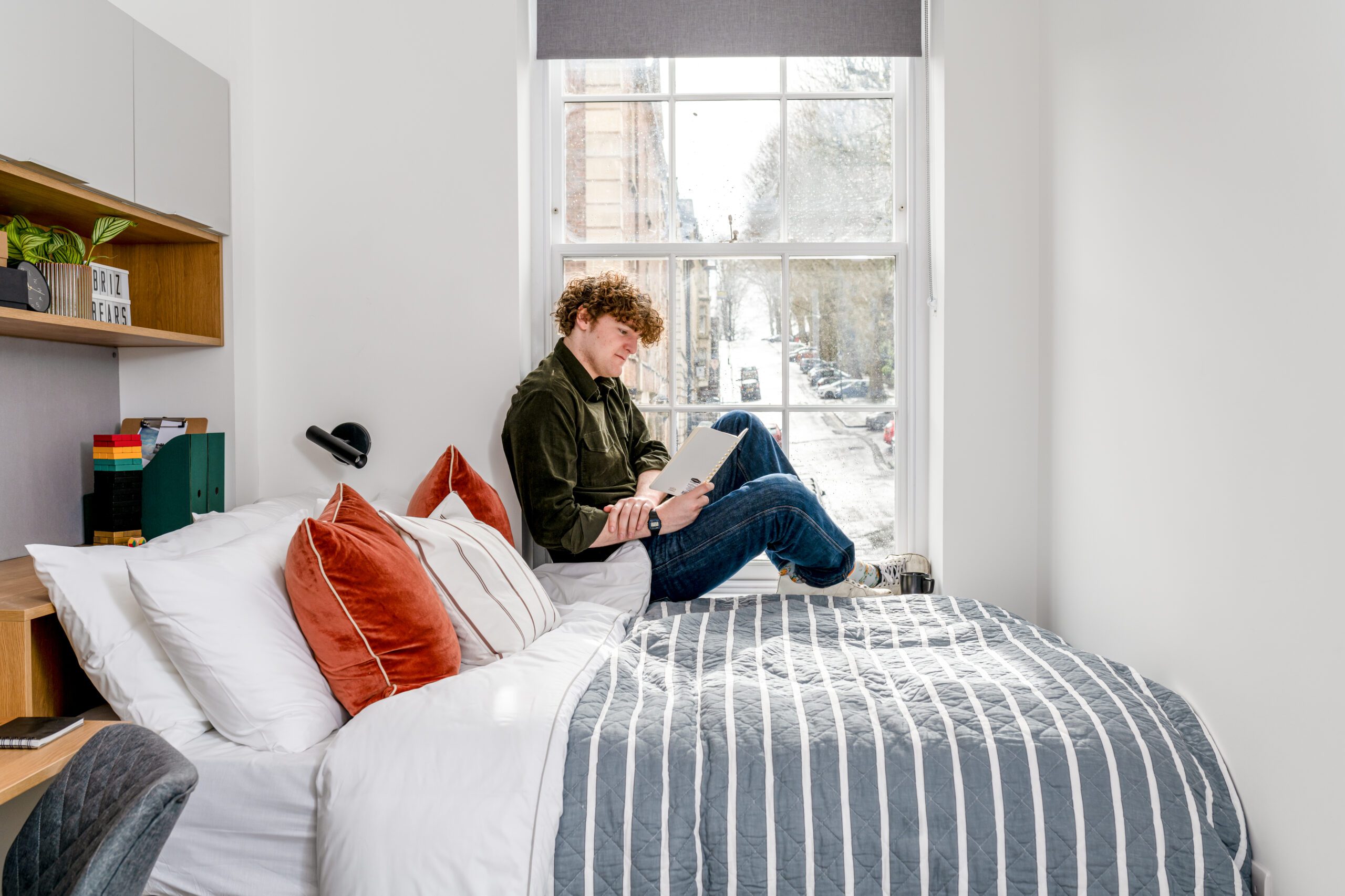 Man sat on window ledge by his bed reading a book