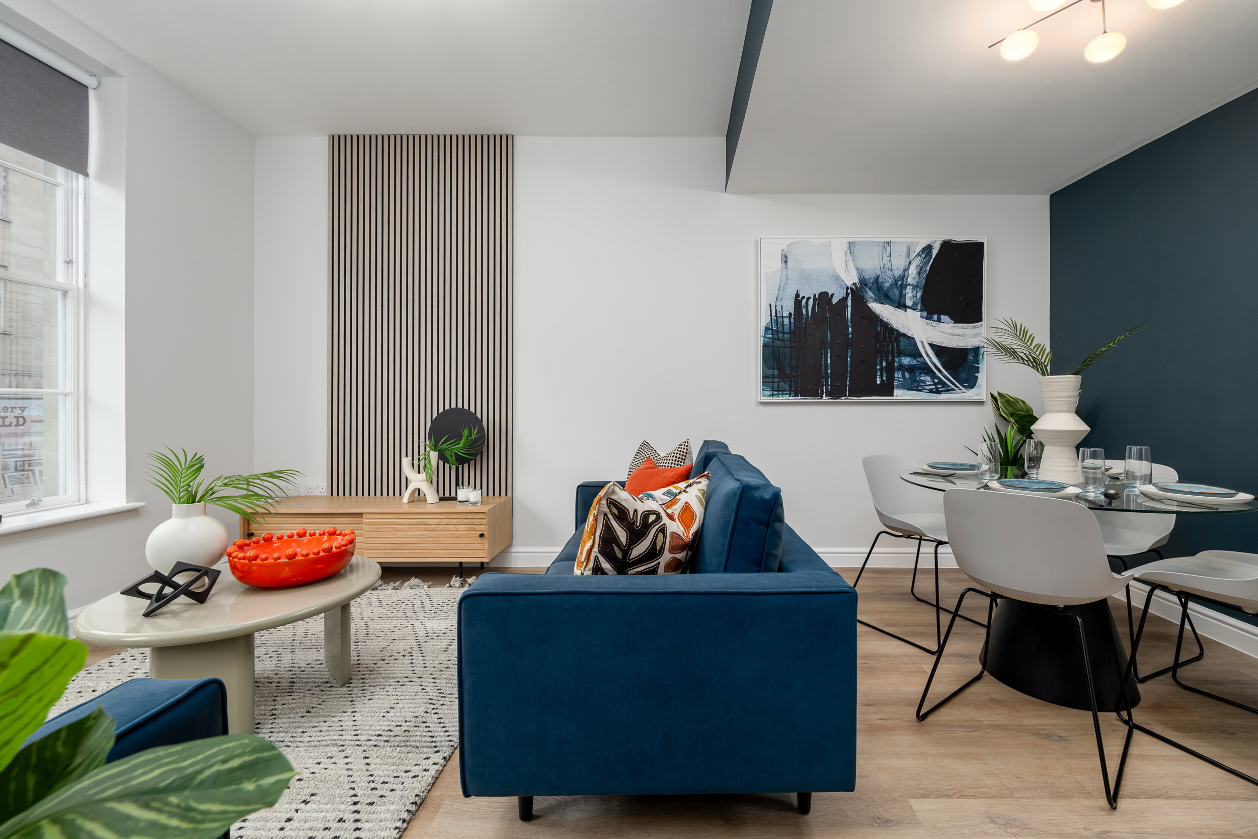 Side shot of living area with blue sofa, glass dining table and ornaments, feature wall and coffee table.