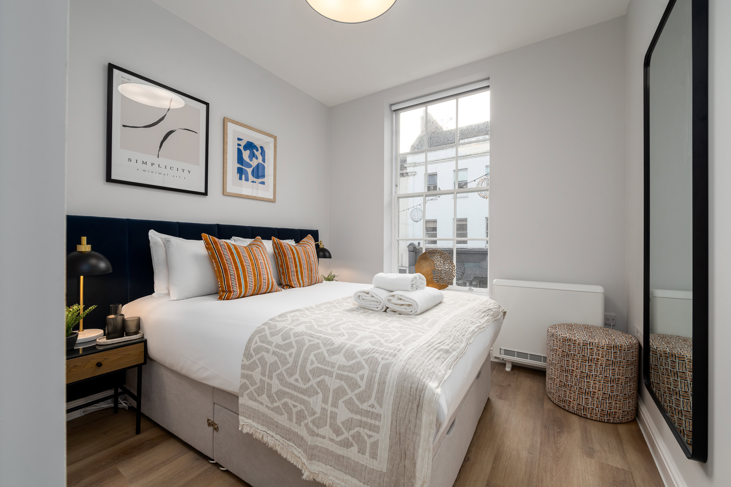 Shot of bedroom with well presented bed with white linen, throw on the end of the bed and yellow cushions. Artwork above and window to the rear.