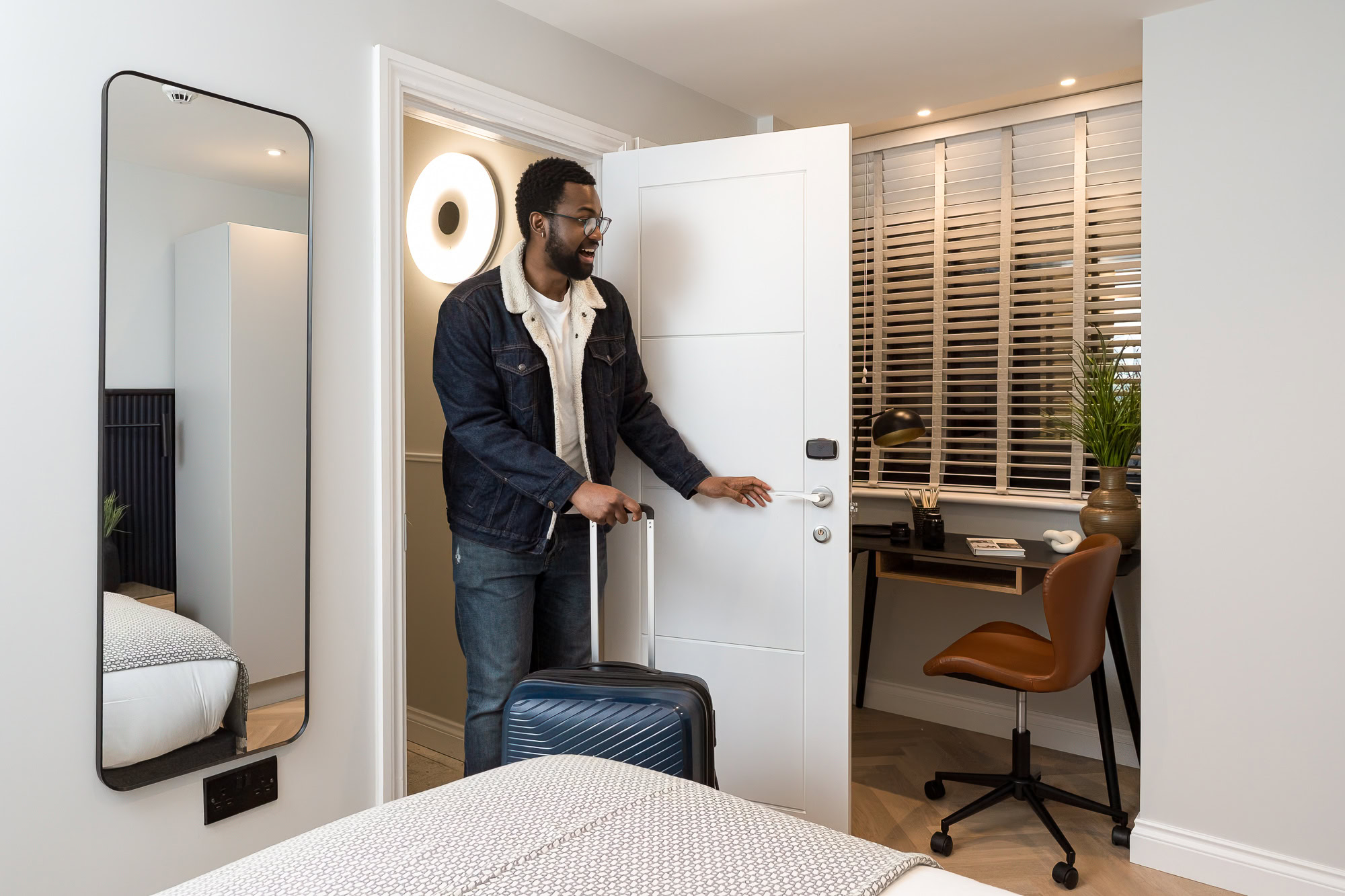 A man stands in front of a door, holding a suitcase, ready to leave or enter a building.